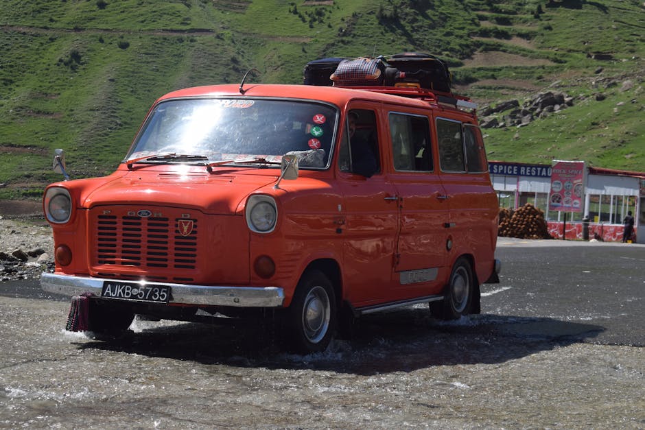 Classic red Ford Transit van crossing a shallow stream in a scenic green landscape.