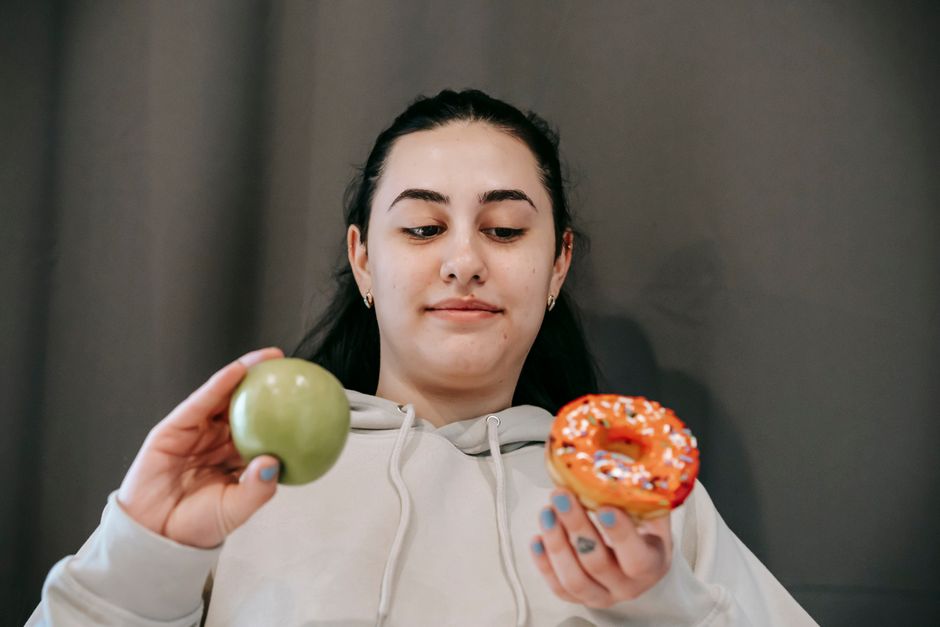 Content young female in hoodie deciding between sweet unhealthy doughnut and green ripe apple in light room