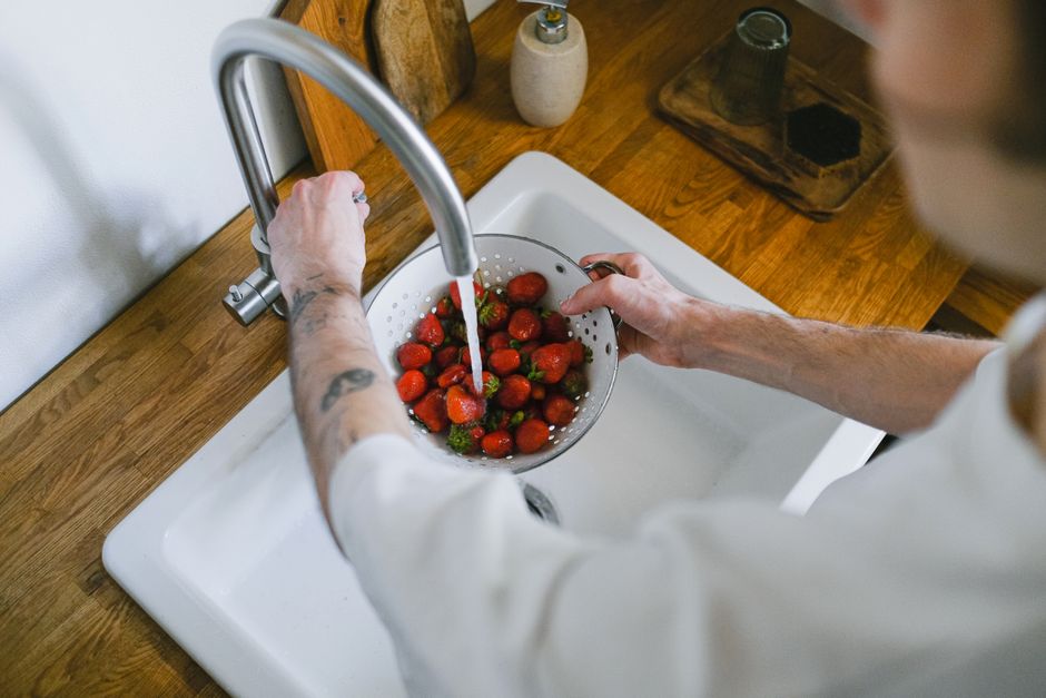 A person washes fresh strawberries in a kitchen sink using a strainer. Overhead angle captures the process.