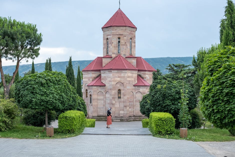 A small stone church with a red roof surrounded by lush greenery and trees.