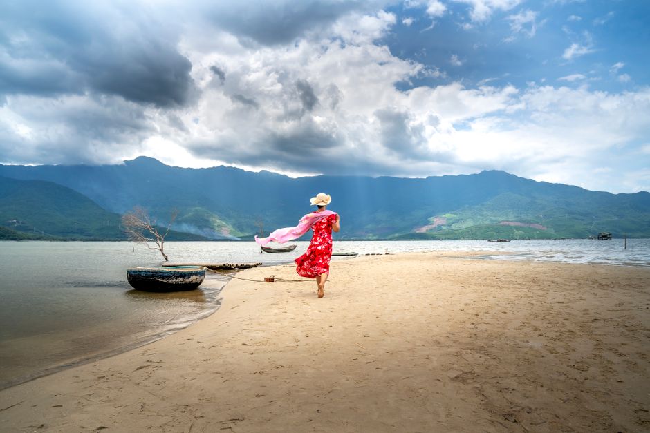 Back view of unrecognizable female traveler with flying textile walking on sandy coast against ocean and ridge under cloudy sky