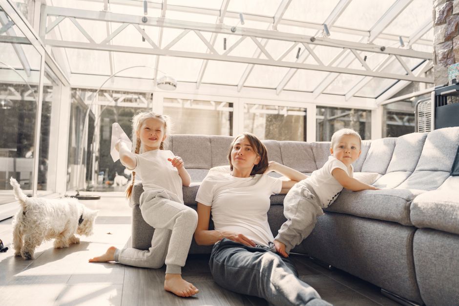 Calm woman sitting on floor and looking at camera while small kids playing around in well lit living room