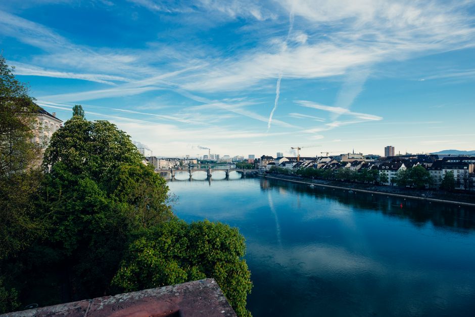 Beautiful view of the Rhine River flowing through Basel, Switzerland, with clear skies and green trees.