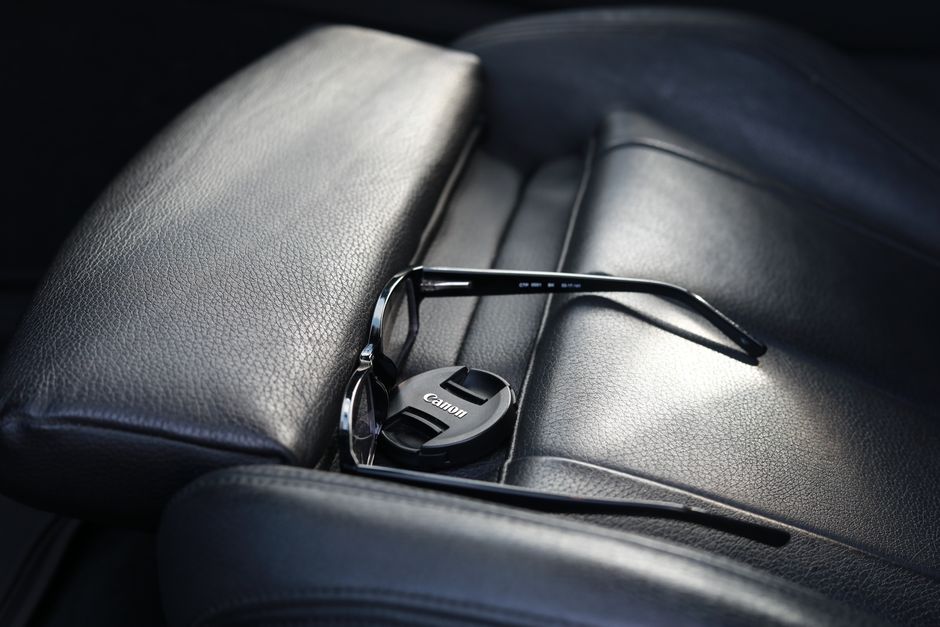 Black sunglasses and camera lens cap resting on a textured leather car seat. Modern and stylish.
