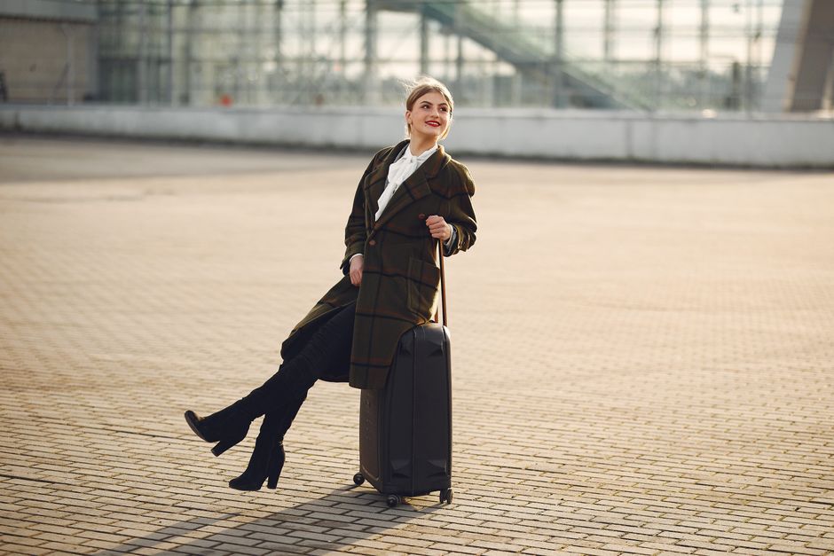 Full length of happy female traveler wearing warm trendy plaid coat and high boots sitting on top of suitcase on pavement near modern airport terminal and smiling looking away during daytime in cold season