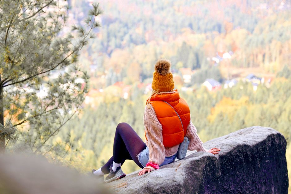 Woman in autumn attire sits on rock, admiring forested mountain view.