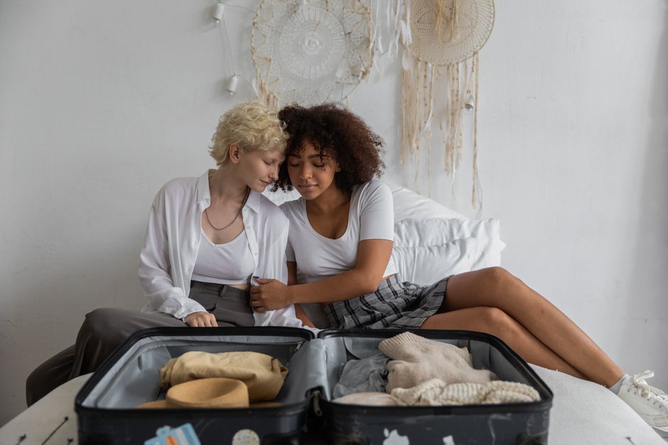Multiracial young girlfriends in casual clothes embracing with closed eyes while packing suitcase in cozy bedroom