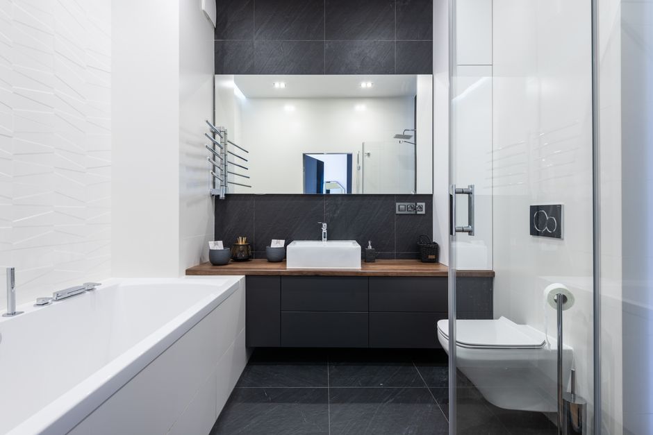 Interior of modern bathroom with bathtub near transparent shower cabin and toilet placed near black cupboard with sink and mirror