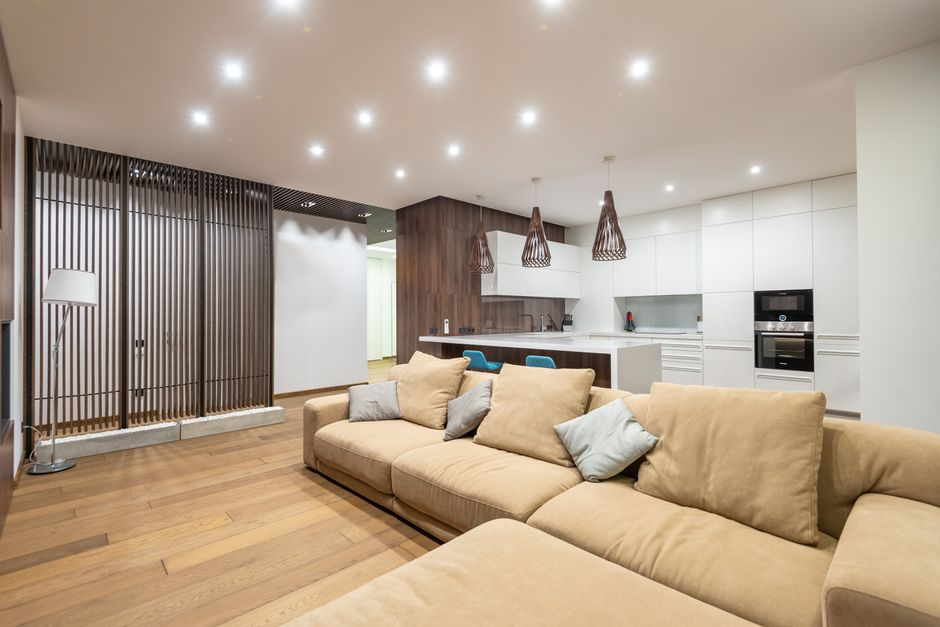 Kitchen zone with white cupboards and modern appliances placed near comfortable sofa with cushions in stylish apartment with wooden decorations